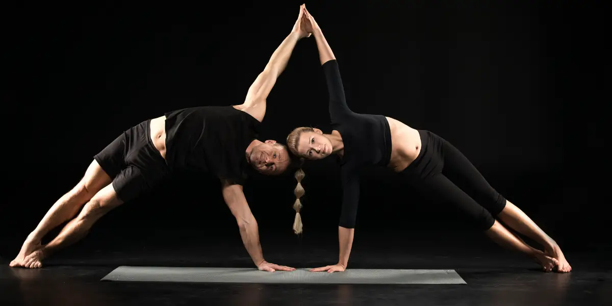 Two people performing a partner yoga pose on a mat against a dark background.