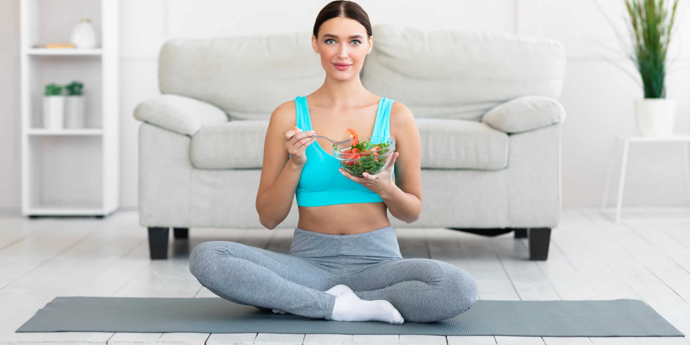 A woman sitting on a yoga mat eating a bowl of salad.