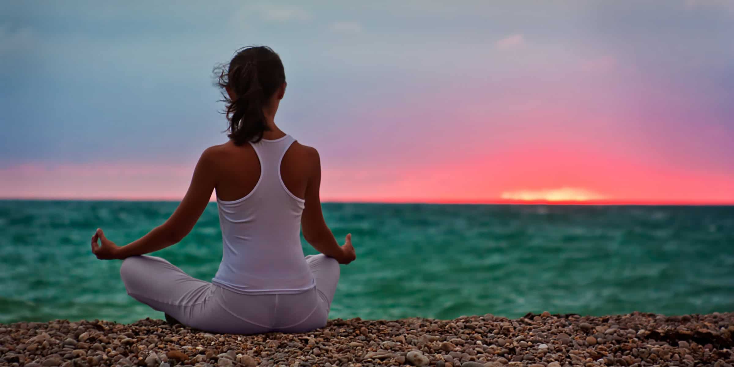 A woman is meditating on the beach at sunset.