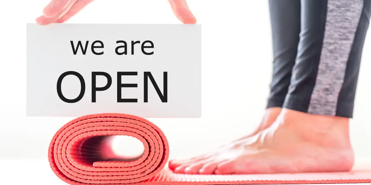 Person standing next to a yoga mat holding a "we are open" sign.