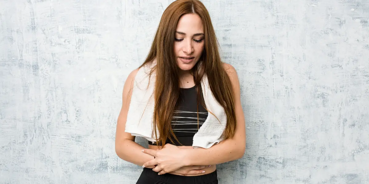 A woman with long brown hair holds her stomach with both hands, wearing a black top and a white towel around her neck, standing against a light textured background.