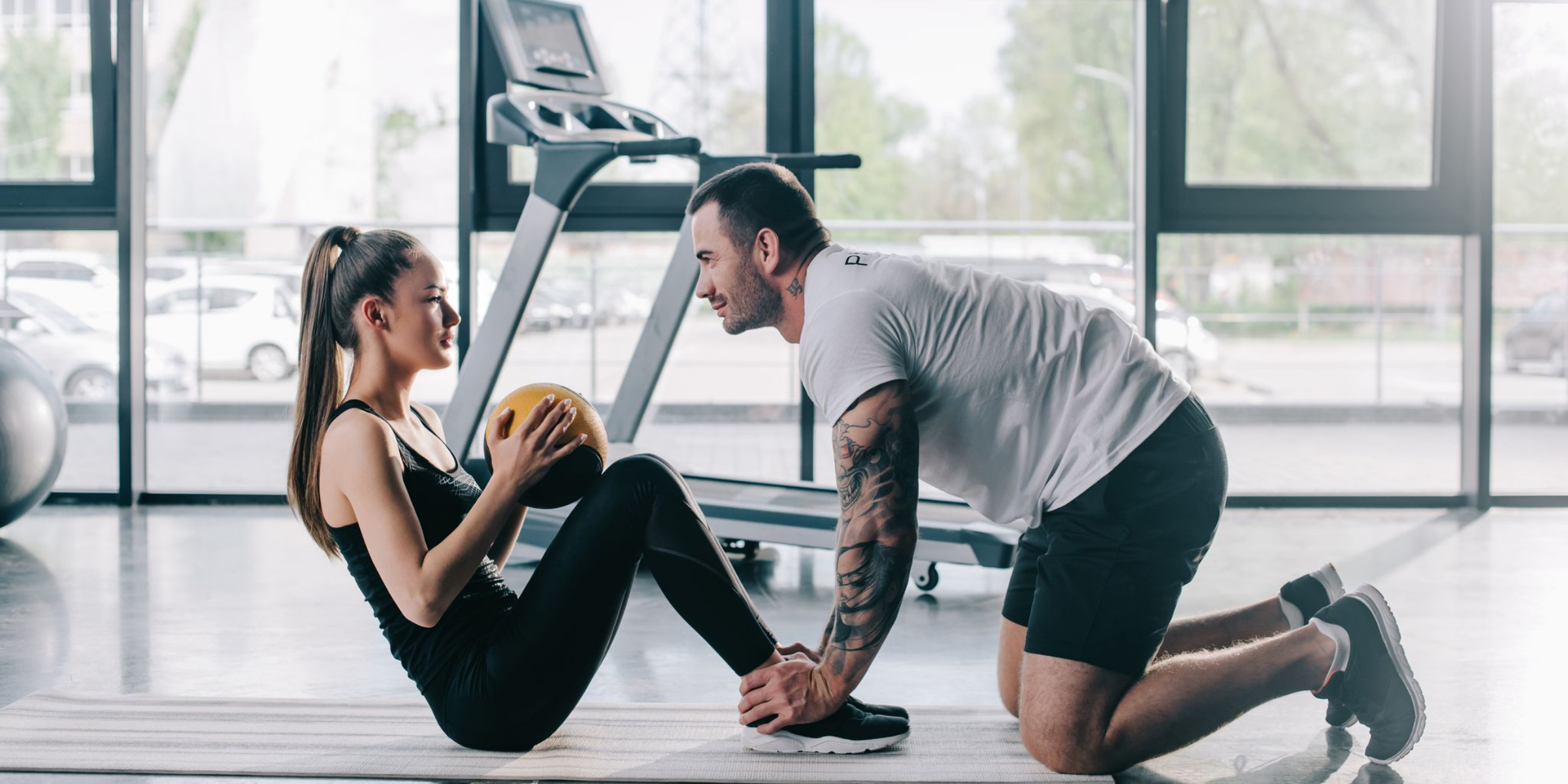 A woman holds a medicine ball while doing sit-ups as a man supports her feet in a gym with a treadmill in the background.